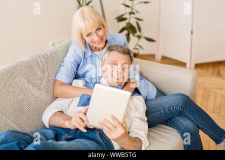 Happy mature spouses using tablet, relaxing on sofa Stock Photo