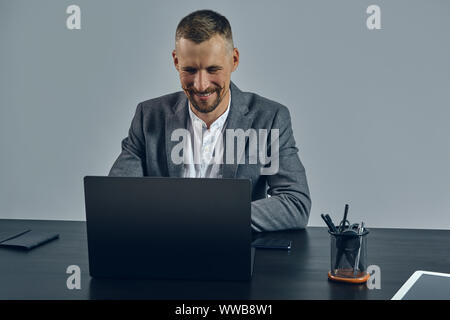 Bearded businessman with stylish mustache, dressed in a classic suit is working at laptop while sitting at table in office, gray background. Stock Photo