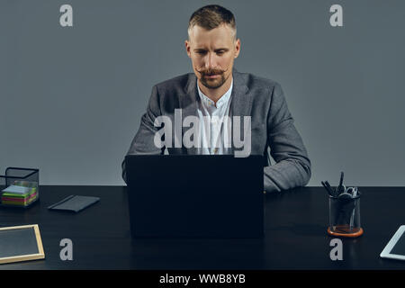 Bearded businessman with stylish mustache, dressed in a classic suit is working at laptop while sitting at table in office, gray background. Stock Photo