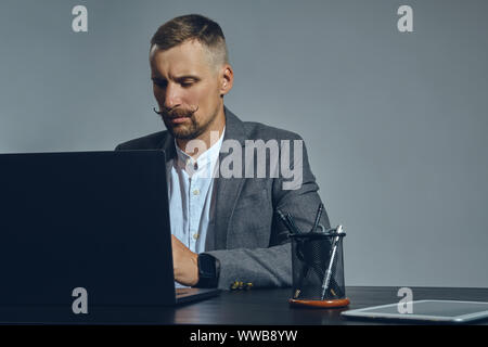 Bearded businessman with stylish mustache, dressed in a classic suit is working at laptop while sitting at table in office, gray background. Stock Photo