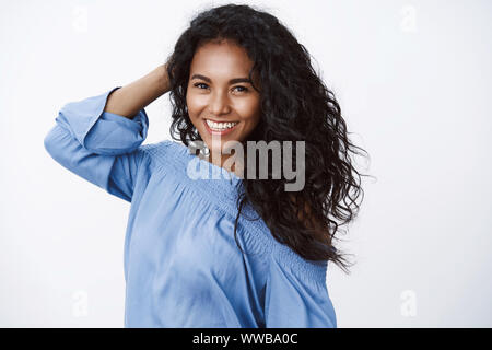 Charming attractive, feminine curly-haired girlfriend in blue stylish blouse, looking fashionably and cheerful, touching hair, flirty gazing camera Stock Photo