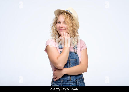 Young curly blonde woman in jeans overalls and summer hat looking confident with smile at camera. Studio shot. Positive facial human emotion Stock Photo