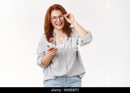 Never was easier. Cheerful carefree happy emotive redhead middle-aged woman checking glasses hold smartphone laughing pleased joyful camera get used Stock Photo