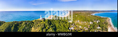 WHite stone Byron Bay lighthouse on the top of green wood covered headland in wide aerial panorama overlooking Byron Bay town and coast with beaches u