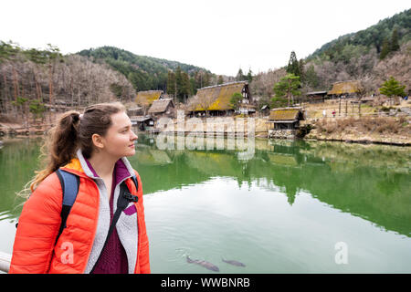 Takayama, Japan with young woman standing by wooden house buildings in traditional Hida no Sato old folk village by green pond lake in early spring Stock Photo