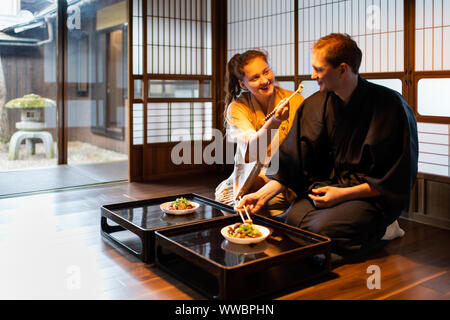 Happy smiling couple in kimono seiza sitting holding chopsticks, eating feeding food at traditional Japanese home room by table plates near shoji slid Stock Photo