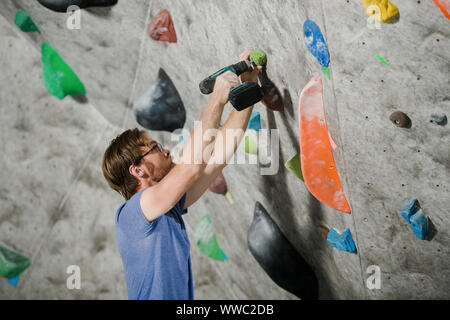 active young bearded man in glasses building climbing wall indoors using drill tool Stock Photo