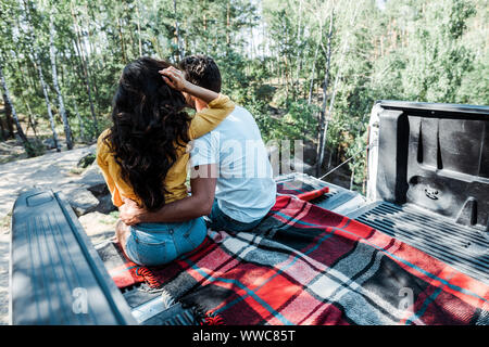 back view of man hugging woman while sitting in car trunk in woods Stock Photo