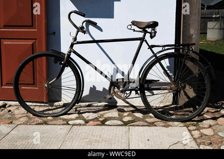 antique black bicycle parked outside old house with wooden door, casting strong shadows on white wall. Ecological means of transport. Stock Photo