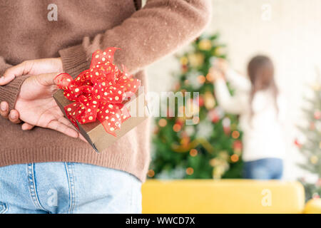 Man hold christmas gift behind his back for surprise his girlfriend while she decorating christmas tree in Chgristmas holiday season greeting. Stock Photo