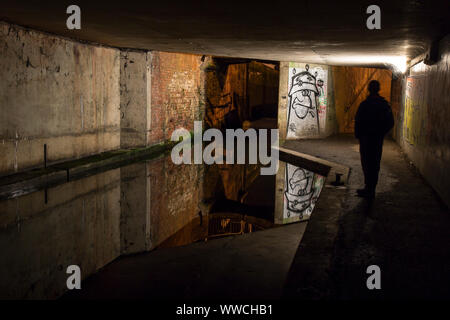 Dark, moody, solitary figure standing by an urban canal in UK pedestrian underpass at night, staring at urban graffiti artwork sprayed on walls Stock Photo