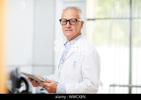 Waist up portrait of senior doctor looking at camera while using digital tablet standing in office of modern clinic, copy space Stock Photo