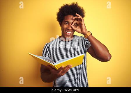 Afro american student man reading book standing over isolated yellow background with happy face smiling doing ok sign with hand on eye looking through Stock Photo