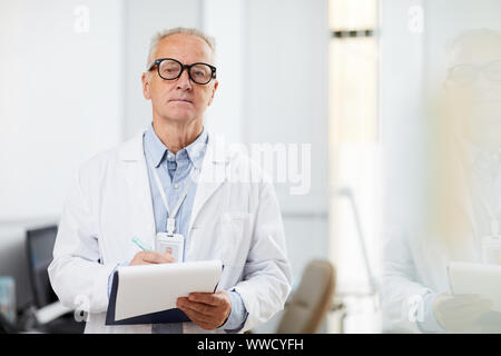 Waist up portrait of senior doctor holding clipboard and looking away pensively while taking notes standing in office, copy space Stock Photo