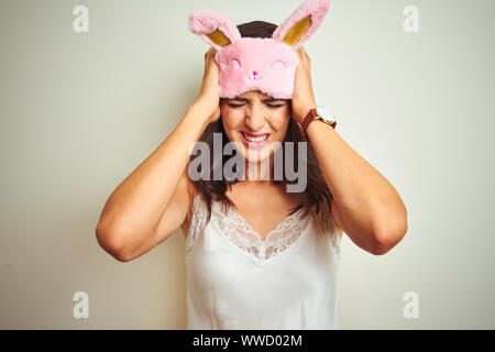 Young beautiful woman wearing pajama and mask standing over white isolated background suffering from headache desperate and stressed because pain and
