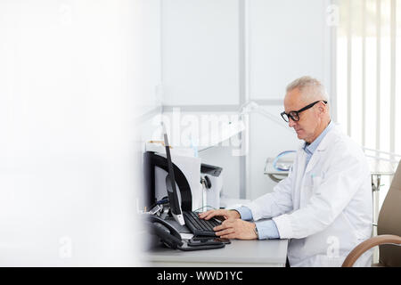 Side view portrait of senior doctor sitting at desk and using computer in office of modern clinic, copy space Stock Photo