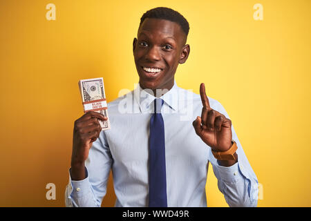 American businessman holding bunch of dollars standing over isolated yellow background surprised with an idea or question pointing finger with happy f Stock Photo