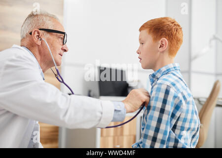 Side view portrait of senior doctor using stethoscope listening to heartbeat and breathing of cute red haired boy during consultation in child healthc Stock Photo