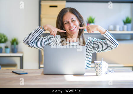Middle age senior woman sitting at the table at home working using computer laptop smiling cheerful showing and pointing with fingers teeth and mouth. Stock Photo