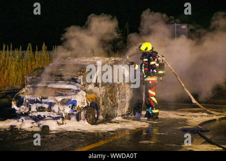 15 September 2019, Germany (German), Oberkrämer: A firefighter stands with a fire hose in front of a burnt-out, smoking vehicle on the A10 motorway at the Oberkrämer junction in the Oberhavel district. In the night to Sunday (15.09.2019) the car was completely burnt out according to first investigations because of a technical defect. (Zu dpa: 'Car burns out on A10 - recovery difficult due to construction site') Photo: Julian Stähle/dpa-Zentralbild/dpa Stock Photo