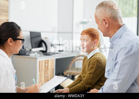 Side view portrait of smiling boy visiting doctor with father and looking at nurse filling patients form in waiting room, copy space Stock Photo