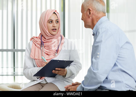 Portrait of young Arab woman working as doctor in medical clinic and talking to senior patient filling in form on clipboard, copy space Stock Photo