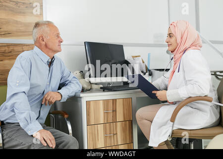 Side view portrait of young Arab woman working as doctor in medical clinic and talking to senior patient filling in form on clipboard, copy space Stock Photo