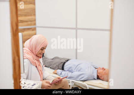 Side view portrait of young Arab woman working as nurse using digital tablet while examining senior patient in medical clinic, copy space Stock Photo
