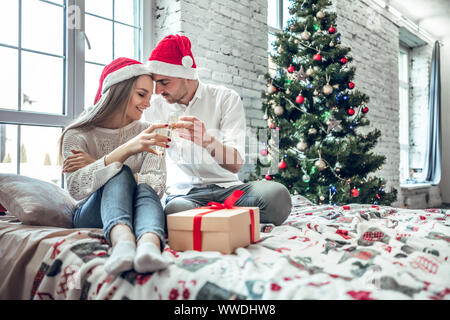 Beautiful couple in love, wearing santa hats, sitting next to a nicely decorated Christmas tree and making a toast with glasses of champagne. Stock Photo