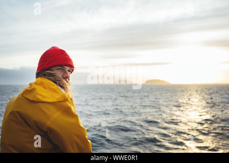 Alone traveler wearing yellow raincoat and red hat looking at sunset sea and foggy mountain. Lifestyle outdoor adventure, scandinavian wanderlust Stock Photo