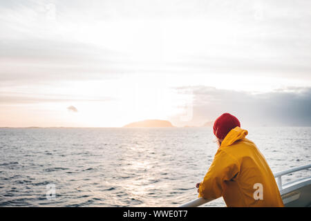 Young traveler wearing red hat and yellow raincoat floating on ship looking at sunset sea after storm and foggy mountains on skyline. Lifestyle travel Stock Photo