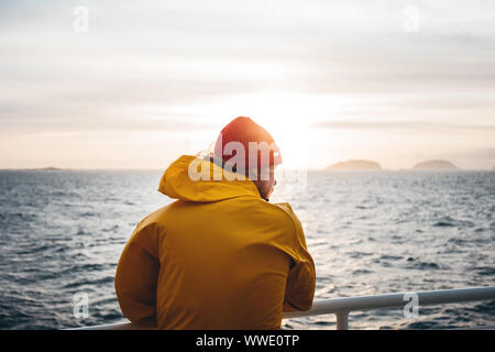 Young traveler wearing red hat and yellow raincoat floating on ship looking at sunset sea after storm and foggy mountains on skyline. Lifestyle travel Stock Photo