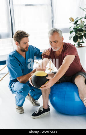 handsome doctor sitting near mature patient exercising on fitness ball Stock Photo