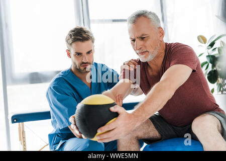 doctor sitting near mature patient exercising on fitness ball Stock Photo