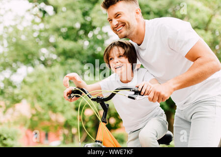 happy father and son looking forward while boy pointing with finger and dad helping kid to ride on bicycle Stock Photo