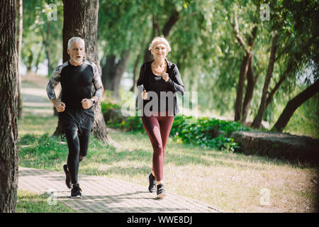 mature, smiling joggers running together on pavement in sunny park Stock Photo