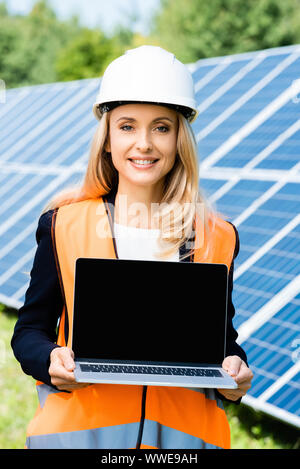 attractive businesswoman in hardhat and safety vest holding laptop with copy space Stock Photo
