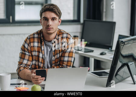 serious young programmer looking at camera while sitting at workplace and holding smartphone Stock Photo