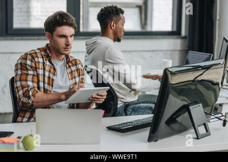 young programmer using digital tablet while sitting near african american colleague Stock Photo