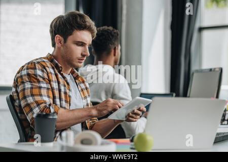 thoughtful programmer using digital tablet while sitting at workplace near african american colleague Stock Photo