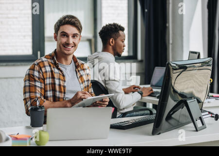 cheerful young programmer smiling at camera while sitting near african american colleague and using digital tablet Stock Photo