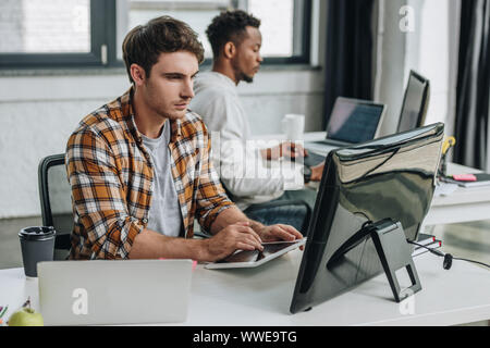 serious young programmer using digital tablet while working in office near african american colleague Stock Photo