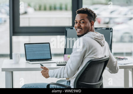 happy african american programmer smiling at camera while sitting at workplace in office Stock Photo