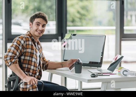 happy programmer smiling at camera while sitting near computer monitor in office Stock Photo
