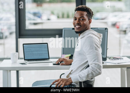 cheerful african american programmer smiling at camera while sitting at workplace in office Stock Photo