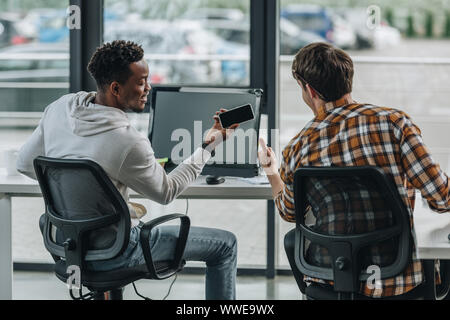 back view of young programmer showing thumb up near african american colleague holding smartphone with blank screen Stock Photo