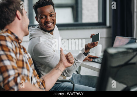 selective focus of smiling african american programmer holding smartphone while sitting near colleague showing thumb up Stock Photo