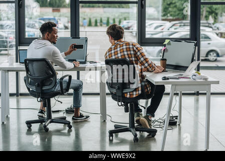 back view of young programmer sitting near african american colleague holding smartphone Stock Photo
