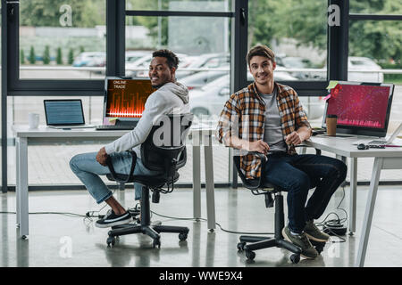 two cheerful multicultural programmers smiling at camera while sitting near computer monitors with graphs and charts on screen Stock Photo