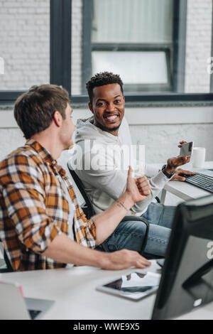 cheerful african american programmer holding smartphone and looking at camera while sitting near colleague showing thumb up Stock Photo
